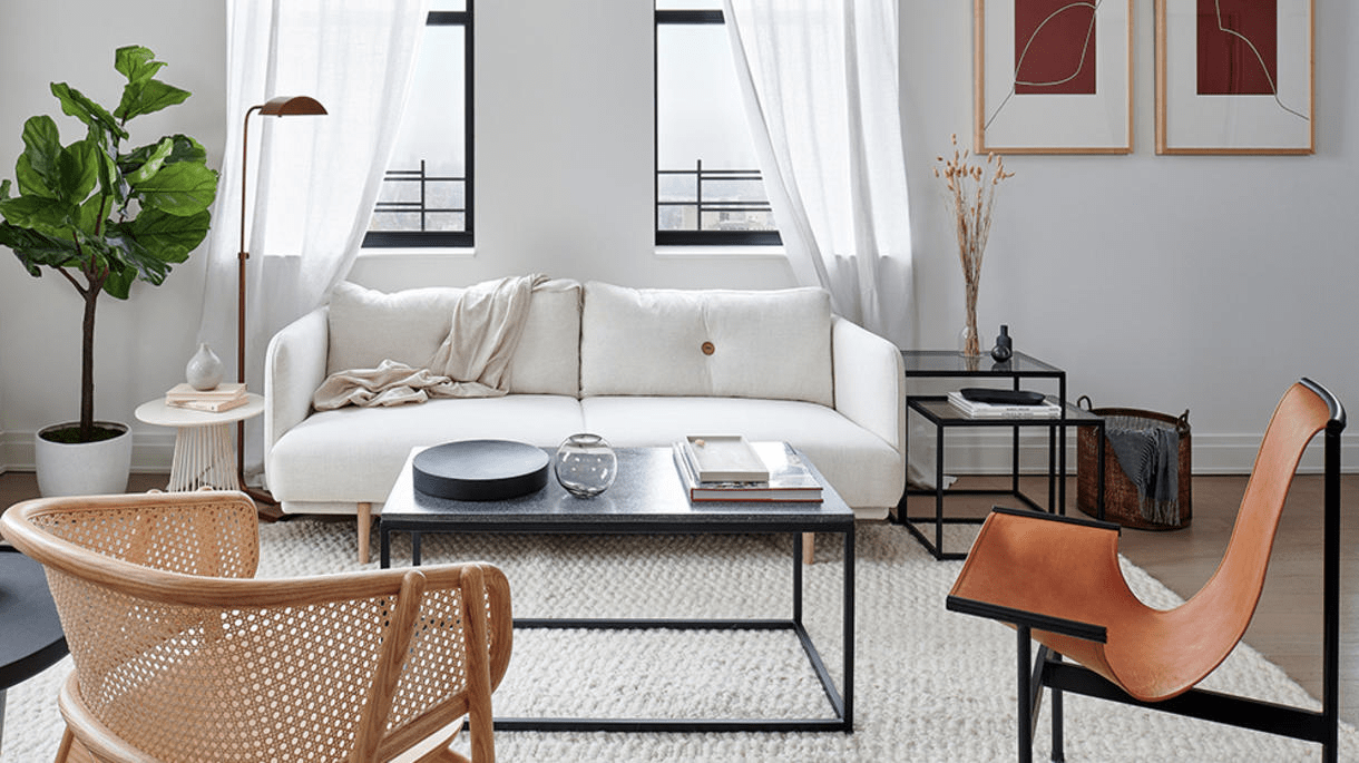 A living room decorated with cream, black, and brown tone furniture, framed by windows and natural light.